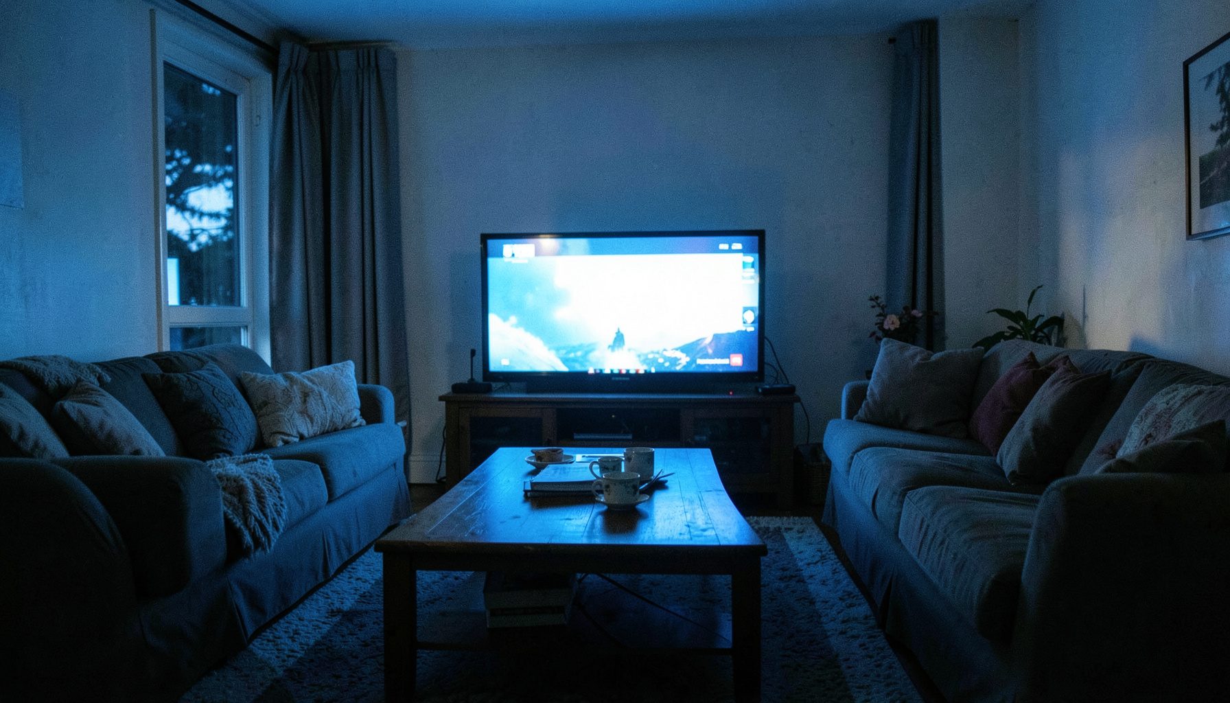 A warmly lit living room at dusk with a television screen glowing in the center, casting soft blue light across a cozy sofa and coffee table