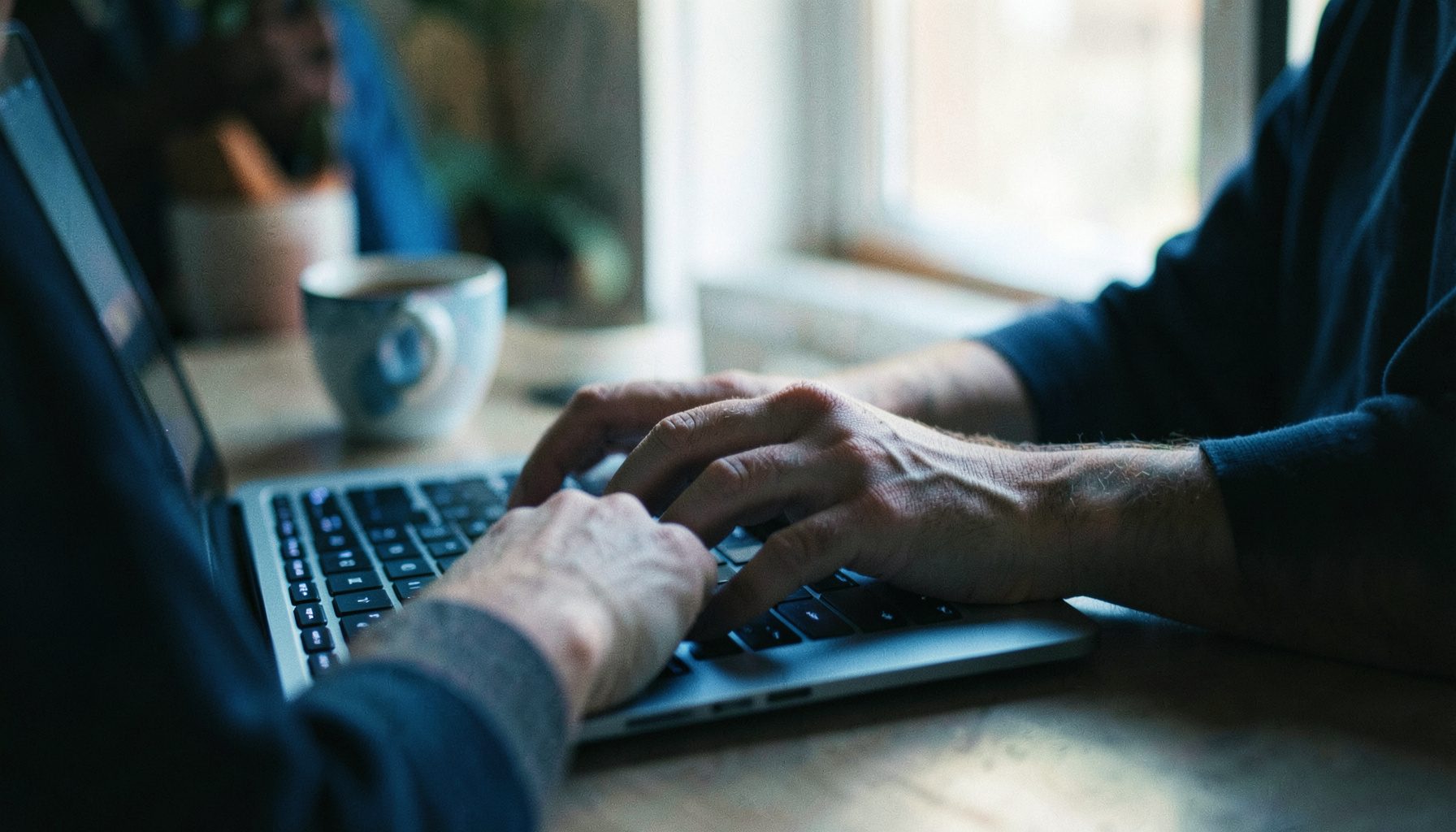 A person's hands typing on a laptop keyboard in a softly lit room, with a mug of coffee nearby and warm ambient light filling the frame.